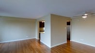 This single-floor space is characterized by a clean, open layout with warm hardwood flooring that extends throughout the main living area, complementing the neutral beige walls and white trim. The living area is generously proportioned, offering a bright and airy feel enhanced by smooth white ceilings.

A partial wall divides the living space from the kitchen, which is accessible through an open archway. The kitchen features dark wood cabinetry, granite countertops, and a stainless steel refrigerator, with overhead pendant lighting providing focused illumination. A breakfast bar or pass-through counter connects the kitchen to the adjoining space, promoting an open and functional flow between areas.

A second opening on the opposite side of the dividing wall leads to an additional room or utility area, also finished with the same hardwood flooring, maintaining visual consistency throughout the space. The overall ambiance is understated and neutral, providing a versatile backdrop well-suited for a variety of furnishing styles. The space presents a practical and cohesive design with attention to material consistency and efficient use of the floor plan.