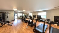 Located at 200 E Mountain St in Worcester, MA, this 670-square-foot fitness center is a fully equipped, single-room facility designed to support a well-rounded workout routine. The space features warm hardwood-style flooring throughout and neutral gray walls, creating a clean and functional environment. Multiple windows along the perimeter allow natural light to fill the room, offering views of the surrounding greenery. The ceiling varies in height with a subtle vaulted section, adding a sense of openness to the compact space. Cardio equipment is well-represented, with a row of TRUE brand treadmills, elliptical machines, and stationary cycling bikes positioned throughout the floor. A multi-station cable strength machine anchors one side of the room, complemented by a dumbbell rack along the wall for free weight training. Wall-mounted yoga mat storage and a water cooler provide added convenience for users. A bulletin board and wall clock are also present, keeping members informed and on schedule. Recessed and flush-mounted ceiling lights ensure ample illumination during all hours. The layout allows for comfortable movement between stations, making this fitness center a practical and well-appointed amenity.
