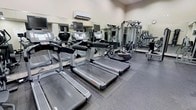 This fitness center is a fully outfitted exercise facility designed to accommodate a wide variety of training needs. The space is anchored by a row of Life Fitness treadmills, each equipped with individual display screens for entertainment and workout tracking, making cardio sessions both effective and engaging. Complementing the treadmills are additional cardio machines, including stationary bikes, providing members with diverse aerobic options. Toward the back of the room, a multi-station strength training machine offers a broad range of resistance exercises, catering to users at all fitness levels. Full-length mirrors line the walls, enhancing the sense of space while allowing users to monitor their form during workouts. The flooring consists of durable rubber matting, providing both safety and comfort underfoot. A wall-mounted air conditioning unit ensures a consistently comfortable climate throughout exercise sessions. The overall layout is thoughtfully organized, with equipment spaced to allow for ease of movement and accessibility. The neutral-toned walls and practical, no-frills design keep the focus firmly on functionality, making this a highly practical and efficient fitness environment.