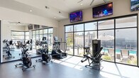 Located at 5959 Cougar Ln in Kearns, Utah, this single-story fitness facility offers a well-appointed exercise space designed to support a comprehensive workout experience. The building's modern exterior features stacked stone and smooth stucco cladding, complemented by low-maintenance desert landscaping and an outdoor patio with seating and a fire feature.

The expansive exercise room is the centerpiece of the facility, bathed in abundant natural light streaming through large, floor-to-ceiling black-framed windows that offer direct views of the outdoor pool and sundeck. The high ceilings, recessed lighting, and oversized ceiling fans contribute to an open, airy atmosphere. The room is equipped with a wide range of TRUE-brand cardio machines — including treadmills, ellipticals, and a stationary bike — alongside a multi-station cable machine, a leg press, free weights, and a dumbbell rack. Wall-mounted flat-screen televisions are positioned throughout for entertainment during workouts, and rubber flooring provides safety and durability underfoot.

The adjacent outdoor pool area features a generously sized swimming pool, lounge chairs, striped umbrellas, and a covered cabana with an outdoor television — creating a seamless indoor-outdoor amenity experience. A connecting hallway links the facility's zones for convenient navigation throughout the space.