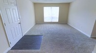 This single-floor space offers a straightforward and functional layout designed to accommodate a variety of living arrangements. The main living area is finished with wall-to-wall carpet in a neutral beige tone, creating a warm and understated foundation that pairs well with a range of furnishing styles. Soft, cream-colored walls throughout contribute to a clean and open atmosphere, while white baseboards provide a crisp contrast that defines the room's boundaries. A large window serves as a focal point of the living area, allowing natural light to fill the space and offering a view of the surrounding exterior. The window's substantial size ensures the room remains bright and airy throughout the day. The open floor plan provides flexibility in how the space can be arranged and utilized. A dark floor panel is visible near the entrance area, adding a subtle material contrast to the otherwise uniform carpeted flooring. The overall ambiance of the space is calm and neutral, offering a blank canvas well-suited for customization and personalization to meet a wide range of needs and preferences.