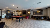 Located at 8 Tice Rd in Franklin Lakes, NJ, this single-floor amenity space is designed with a sleek, upscale aesthetic that blends bold materials with refined furnishings. The open layout is anchored by a full-size billiards table with a rich red felt surface, positioned beneath a striking sculptural chandelier that serves as a visual centerpiece for the room.

Adjacent to the billiards area is a fully equipped kitchen featuring dark, wood-grain cabinetry, a stainless steel refrigerator, reflective black glass backsplash, and a dramatic oversized island clad in deep black stone with bold gold veining. Gold pendant chandeliers with a tiered, architectural design illuminate the space with warm, ambient light.

A dedicated lounge zone anchors one end of the space, where a floor-to-ceiling dark stone accent wall houses a wall-mounted flat-screen TV above a linear electric fireplace with gold flame elements. Vibrant magenta velvet accent chairs and gold-accented side tables add a pop of color against the moody backdrop.

A dining nook features a sleek black oval table surrounded by modern chairs with gold bases, positioned beneath a large gold drum pendant. Textured wall art and sconce lighting contribute to the layered, upscale atmosphere throughout.

A secondary lounge area offers plush dark sofas, tufted accent chairs, and patterned area rugs over light wood-look flooring, creating an inviting, comfortable gathering space. The overall ambiance is modern, glamorous, and highly functional.