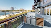 Located at 25 Lewis St in East Boston's vibrant waterfront district, this outdoor amenity terrace offers a compelling blend of modern design and breathtaking natural scenery. The space is defined by its expansive deck, combining composite decking with tasteful stone paver sections that create distinct zones for relaxation and entertaining. Floor-to-ceiling glass doors and large windows along the building façade connect the terrace seamlessly to the interior, while a sleek brick and panel exterior wall adds architectural texture and depth.

The terrace is outfitted with a stainless steel Weber gas grill station, elegantly integrated into custom wood-slatted cabinetry, making it ideal for outdoor cooking and social gatherings. Multiple seating arrangements, including white outdoor chairs and benches, are thoughtfully positioned to take advantage of the sweeping panoramic views of Boston Harbor and the downtown Boston skyline. Cable-mesh railings with rich wood-capped handrails ensure unobstructed sightlines across the water.

The waterfront promenade and pier are visible just beyond the terrace, enhancing the sense of openness and connection to the harbor. The overall ambiance is polished and contemporary, offering residents a well-appointed outdoor retreat with direct access to one of Boston's most scenic urban waterfronts.