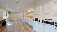Located at 593 Salem Woods Dr SE in Marietta, Georgia, this single-floor shared laundry facility is designed to efficiently serve its users with a clean, functional layout. The space is finished with light wood-style plank flooring throughout, lending a modern, well-maintained aesthetic that extends across the entire room.

The laundry area is equipped with multiple commercial-grade top-loading washing machines and stacked dryer units arranged along the walls, providing ample capacity for shared use. A built-in wooden folding counter runs along one wall, offering a convenient surface for sorting and folding laundry. Waste bins are positioned nearby for easy cleanup. A posted laundry rules sign keeps users informed of facility guidelines.

Recessed lighting illuminates the space evenly from above, complemented by large windows that allow natural light to filter in and provide views of the surrounding wooded landscape. A secondary door offers additional access and further connects the interior to the greenery outside.

The neutral wall tones and clean finishes create a bright, orderly environment. The open floor plan allows for comfortable movement between machines, and the overall design prioritizes practicality while maintaining a tidy, inviting atmosphere suitable for everyday use.