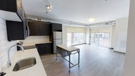 This single-floor space showcases a contemporary open-concept layout that seamlessly integrates the kitchen and living areas. The kitchen is outfitted with dark espresso cabinetry, a classic subway tile backsplash, and stainless steel appliances, including a full-sized refrigerator. Light quartz-style countertops provide a striking contrast against the darker cabinet tones. A mobile island with a metal frame adds flexible workspace and can be repositioned as needed. Exposed ductwork runs along the upper perimeter of the space, lending an industrial character that complements the modern aesthetic. Track lighting illuminates the kitchen area, while a flush fixture brightens the living zone. Wide-plank hardwood-style flooring flows continuously throughout, unifying the open floor plan. Large windows along the exterior wall flood the living area with natural light and offer views of the surrounding streetscape. Roller shades provide adjustable light control. The overall ambiance is clean, modern, and functional, balancing industrial design elements with contemporary finishes.