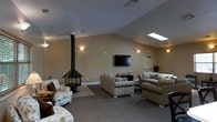 This single-story lounge space is defined by its vaulted ceiling, which lends a sense of openness and airiness to the room. A skylight positioned along the roofline draws in natural light, complementing the warm-toned walls and neutral carpeted flooring. A striking freestanding black wood-burning stove serves as a focal point, its tall cylindrical flue pipe extending toward the ceiling and adding a rustic, cozy character to the space.

The seating arrangement includes multiple plush armchairs and a sofa upholstered in classic patterned fabrics, thoughtfully grouped around a central area rug to create an intimate gathering environment. A wall-mounted flat-screen television is positioned at a comfortable viewing height, supported by a low media console beneath it. Soft wall sconces provide ambient lighting throughout the room, enhancing the welcoming atmosphere.

A doorway visible toward the back of the space suggests additional rooms or access points beyond, while a touch of greenery near the entrance adds a natural accent. Overall, the space is well-suited as a shared relaxation or social lounge area.
