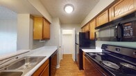 This single-floor residence offers a well-organized and functional layout across its living spaces. The galley-style kitchen is efficiently designed with warm honey-toned wood cabinetry lining both walls, light speckled countertops, a stainless steel sink, and a full suite of black appliances including a refrigerator, electric range, over-the-range microwave, and dishwasher. Hardwood-style flooring runs throughout the kitchen, and a walk-in closet or pantry is conveniently accessible directly from the kitchen area. A textured ceiling and ceiling-mounted light fixture complete the kitchen's practical design. The living area and bedroom feature neutral-toned carpet underfoot and textured ceilings, contributing to a warm, comfortable ambiance. A ceiling fan with a light kit is installed in the bedroom, which also benefits from a window that invites natural light. The main living area is highlighted by a full-width sliding glass door fitted with vertical blinds, offering views of lush greenery outside. The hallway connects the various spaces seamlessly, with doors leading to additional rooms. The bathroom is clean and straightforward, featuring a single vanity with speckled countertop, wood cabinetry below, a large mirror with overhead lighting, and a full tub-shower combination with built-in shelving.