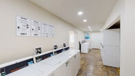 This single-story shared laundry facility in Gretna, Louisiana, is designed to accommodate a high volume of use with a practical, functional layout. The space is organized into two connected areas, each serving a distinct purpose within the laundry operation.

The main laundry area features a row of commercial-grade top-load washing machines lining one wall, accompanied by stacked dryer units along the opposite side. Digital controls on each machine allow for straightforward operation, and informational signage — including laundry tips and service instructions in both English and Spanish — is posted prominently for user guidance. A dedicated CSC payment system is integrated into the equipment, supporting modern cashless transactions.

The interior is finished with neutral, light-toned walls and recessed ceiling lighting that keeps the space well-illuminated throughout. Multiple windows along the walls invite natural light and offer views of the surrounding greenery, contributing to an open and airy atmosphere. A wall-mounted ventilation fan provides additional airflow, which is especially beneficial in the humid Louisiana climate.

The flooring displays a raw, textured concrete surface with varied natural toning throughout both sections of the space. Entry is provided through a solid paneled door, and the overall layout allows for comfortable movement between machines, making this a well-equipped, accessible shared laundry facility.