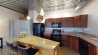 This single-floor loft at Artek Lofts showcases a distinctive industrial aesthetic defined by exposed metal corrugated ceilings with visible ductwork and track lighting throughout much of the unit. The open layout connects key living areas seamlessly, creating a spacious and cohesive flow.

The kitchen is a functional centerpiece, equipped with rich wood cabinetry, black appliances including an electric range, microwave, and refrigerator, and a generous granite-topped island with bar seating. Pendant lights hang above the island, enhancing both style and utility.

The living area features multiple windows fitted with horizontal blinds, flooding the space with natural light. Polished terrazzo-style flooring grounds the main areas, while carpet adds warmth to select rooms.

The bedrooms offer a calm retreat; one notably features a curved wooden ceiling detail that adds architectural interest, along with large windows providing outdoor views. Both bedrooms offer ample space and understated design.

The bathroom presents a clean, classic aesthetic with white subway tile wainscoting, white floor tile, a pedestal sink, and a mirrored vanity. A dedicated laundry area with a stacked washer and dryer and built-in shelving adds practical convenience to this well-appointed loft.