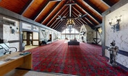 Located at 3710 Inverrary Dr in Lauderhill, Florida, this single-floor Tudor-style clubhouse offers a generously sized, character-rich space designed for community use. The centerpiece of the building is its dramatic great hall, defined by soaring exposed wood beam vaulted ceilings with decorative wrought-iron chandeliers and ceiling fans suspended overhead. Bold, ornate red carpeting runs throughout much of the space, anchoring the warm, lodge-like ambiance. The walls feature distinctive Tudor half-timbering with textured white paneling, lending the interior an unmistakable architectural identity.

A grand floor-to-ceiling stone fireplace with dual openings serves as a stunning focal point at one end of the great hall, complemented by hardwood flooring in the immediate seating area and large glass sliding doors that provide abundant natural light and direct views of the outdoor pool area. The entrance foyer welcomes visitors through double wood doors flanked by colorful diamond-pattern stained glass sidelights, opening into the main hall.

A library room houses extensive built-in wooden bookshelves stocked with reading materials and includes a mounted television, offering a quieter, more intimate setting. A dedicated office and a hallway corridor with tile flooring complete the layout, rounding out a versatile and well-appointed community facility.