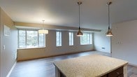 This single-floor space showcases an open-concept layout that seamlessly connects the kitchen and living areas, creating a spacious, airy atmosphere. The kitchen features a generously sized island with a granite or stone-look countertop, providing ample workspace and a natural gathering point. Two stylish pendant lights hang above — one a cylindrical drum shade and the other a sleek metallic drop pendant — adding visual interest and targeted illumination.

The living area extends comfortably beyond, finished with smooth hardwood-style flooring in a light, neutral tone that enhances the sense of openness throughout. A structural beam detail runs along the ceiling, adding architectural character while maintaining the room's clean, contemporary aesthetic.

A row of large windows lines the far wall, flooding the space with natural light and offering pleasant views of surrounding greenery. The neutral wall tones complement the flooring and trim, creating a cohesive, understated palette. Recessed ventilation and subtle baseboards complete the well-finished interior. Overall, the space conveys a modern, functional design with an emphasis on light, flow, and clean architectural detail.