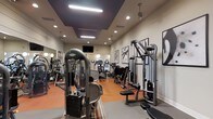 This fitness center offers a thoughtfully designed workout space equipped with a comprehensive selection of exercise machines. The room features a variety of strength-training equipment, including cable machines, weight stacks, and resistance apparatus, alongside cardio machines such as ellipticals and upright bikes arranged toward the rear of the space. The flooring combines durable orange and gray tones, delineating workout zones while adding a modern visual contrast. Overhead, a coffered ceiling detail with recessed lighting and pendant fixtures provides bright, even illumination throughout the space. A wall-mounted flat-screen television is positioned for easy viewing during workouts. Large framed artwork with bold graphic designs lines the walls, contributing an elevated aesthetic beyond a typical gym environment. Mirrors along one wall enhance the sense of openness and allow users to monitor their form. The neutral, warm wall tones paired with the contemporary ceiling design give the space a polished, welcoming ambiance suitable for a range of fitness activities.