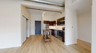Located at 135 CTH-125 in Appleton, Wisconsin, this single-floor unit showcases a contemporary industrial aesthetic blended with modern finishes throughout. The open-concept living and kitchen area is anchored by light hardwood-style flooring, soaring ceilings with exposed spiral ductwork, and black rail accents that lend an urban, loft-inspired character to the space. A sliding glass door leads to an outdoor balcony, allowing natural light to flood the generous living area.

The kitchen is well-appointed with dark espresso cabinetry, a full suite of stainless steel appliances including a refrigerator, electric range, built-in microwave, and dishwasher, complemented by light countertops and a single-basin sink. A rustic wood island provides additional prep and gathering space.

The bedroom features plush grey carpeting, a large window with exterior views, and a sliding barn-style door that opens to a spacious walk-in closet with built-in shelving. The bathroom is finished with large-format tile flooring, a dark wood vanity with a white countertop, a well-lit mirror, and a walk-in shower with built-in shelving and grab bars. The unit's clean, neutral palette and thoughtful layout make it a highly functional and visually cohesive living space.