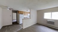 Located at 221 South Oak Avenue in Clifton Heights, Pennsylvania, this single-floor residence offers a functional and straightforward layout designed for comfortable everyday living. The kitchen is positioned at the heart of the unit, featuring a peninsula-style counter that separates it from the main living area while maintaining an open, connected feel. Light wood cabinetry provides ample storage, and white appliances — including a full-size refrigerator, microwave, and range — are integrated neatly into the space. A built-in sink is set into the peninsula counter, adding to the kitchen's practicality. The living area extends openly from the kitchen, finished with wall-to-wall carpeting that lends a warm, understated ambiance. A window along the side wall allows natural light to filter into the space. A hallway visible from the main living area leads to additional rooms, suggesting a well-organized floor plan. The walls are finished in a neutral tone throughout, creating a clean and versatile backdrop. Baseboard heating units are installed along the walls, ensuring consistent warmth. The textured ceiling adds subtle dimension to the interior without detracting from the overall simplicity of the design.