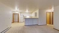 This single-floor residence offers a practical and well-organized open-concept layout that seamlessly connects the kitchen, dining, and living areas into one cohesive space. The kitchen is equipped with ample white cabinetry providing generous storage, a white refrigerator, and a peninsula-style countertop that serves as a natural divider between the kitchen and the living area. The peninsula also functions as a convenient breakfast bar or prep space, adding to the room's versatility. Light-toned carpeting extends throughout the living area, creating a warm and neutral foundation that complements the clean white walls and cabinetry. A ceiling fan with integrated lighting is centrally positioned, ensuring comfortable air circulation and adequate illumination throughout the open space. A walk-in closet fitted with built-in shelving and organizational components offers excellent storage capacity. The overall design is straightforward and functional, with a neutral color palette of whites and soft beiges that creates a bright, clean atmosphere throughout. The layout is well-suited for efficient everyday living, with each area flowing naturally into the next.