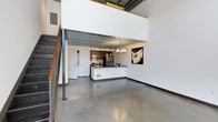 This two-story loft showcases a striking blend of industrial and modern design elements. The main level features polished concrete floors that extend throughout the open-concept living and kitchen area, lending the space a sleek, contemporary feel. Soaring double-height ceilings amplify the sense of volume, while exposed corrugated metal ceiling details and structural elements reinforce the industrial character of the space.

The kitchen is centrally positioned and features dark wood cabinetry, stainless steel appliances including a refrigerator and microwave, and a peninsula with a dark countertop that provides both prep space and a casual dining surface. Track lighting above the kitchen is complemented by a chandelier, offering layered illumination. A staircase with metal railings and warm wood treads leads to the upper mezzanine level, which overlooks the main living area below and features greenery along its railing. The open sightlines between levels enhance the airy, expansive feel of the space. Overall, the loft presents a cohesive, contemporary industrial ambiance.