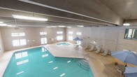 This single-floor indoor recreational facility is designed to provide a comprehensive aquatic and leisure experience. The centerpiece of the space is a generously sized indoor swimming pool with clear turquoise water, featuring entry steps with railings for easy access. Adjacent to the main pool sits a circular hot tub, providing a dedicated space for relaxation. A poolside patio umbrella shades a small table and chairs, while a row of lounge chairs lines the surrounding deck, offering ample space for guests to rest and unwind. Safety equipment, including a life ring, is visibly mounted on the wall. The pool area is enclosed by neutral-toned masonry block walls and illuminated by rows of overhead fluorescent lighting. A series of windows along the upper portion of the walls allows natural light to filter into the space, enhancing the bright and open atmosphere. A secondary lounge area is also part of the facility, featuring multiple tables and chairs arranged on dark carpeted flooring beneath high ceilings with industrial-style overhead lighting. This area is framed by the same masonry block walls and provides a comfortable gathering space separate from the pool deck. Access doors connect the various areas of the facility, ensuring convenient circulation throughout.