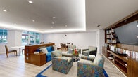 Located at 410 Evernia Street in West Palm Beach, Florida, this well-appointed game room offers a thoughtfully designed, single-floor layout that balances comfort and functionality. The space is finished with light wood-look flooring throughout and features a striking tray ceiling with integrated ambient lighting that lends a modern, polished look to the interior.

The central lounge area serves as the focal point, anchored by a set of colorful, patterned armchairs arranged around a glass coffee table atop a blue-bordered area rug. A built-in warm wood shelving wall houses an extensive book and media collection alongside a large flat-screen television, creating an inviting media and library nook. Adjacent to the lounge, a vivid blue sofa with accent pillows adds a bold splash of color.

A separate dining area provides multiple white-topped tables with upholstered wood-frame chairs. Large glass doors open directly to an outdoor patio, flooding the dining zone with natural light and views of lush greenery. A service kitchenette with warm wood cabinetry and a pass-through window connects conveniently to the main lounge area. A connecting corridor with light grey walls and clean white trim completes the layout, providing seamless circulation throughout the space.