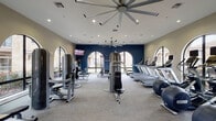 This fitness center in Spring, TX offers a generously sized, single-floor workout facility designed to support a comprehensive exercise routine. The space is characterized by its striking architectural detail — a row of tall, arched windows lines the walls, flooding the interior with natural light and providing views of the surrounding landscaped grounds. A bold navy blue accent wall adds a modern, energetic contrast to the neutral beige tones throughout the rest of the space.

The main workout floor is well-organized and thoughtfully equipped. A full lineup of treadmills and elliptical machines is positioned along the windowed wall, each unit accompanied by an individual screen for entertainment during cardio sessions. Strength training is well-represented with multiple selectorized cable and weight machines, a dumbbell rack with an adjustable bench, and additional resistance equipment spread across the floor.

Overhead, large industrial-style ceiling fans with wide blade spans ensure consistent airflow throughout the space. Recessed lighting provides even, bright illumination. Mounted televisions are positioned for visibility from multiple workout zones.

Entry to the fitness floor is through a set of glass-paneled French doors, with a small cubby storage unit nearby for personal belongings. The flooring transitions from hardwood in the entryway to low-pile carpet in the main exercise area, keeping the space clean and functional.