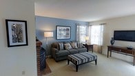 This single-floor residence offers a thoughtfully designed living space that balances comfort and style. The living room serves as the heart of the home, immediately drawing attention with its striking blue accent wall, which provides a rich contrast against the neutral, light-toned walls and ceiling throughout the space. A classic brick fireplace anchors one side of the room, adding warmth and a timeless architectural character to the overall aesthetic.

The living room is generously sized, accommodating a plush sofa adorned with decorative pillows and a coordinating striped ottoman that doubles as a functional centerpiece. A framed piece of artwork hangs prominently above the sofa, complementing the room's understated yet curated décor. Soft carpeting underfoot enhances the cozy atmosphere throughout.

Natural light fills the space through multiple windows dressed with sheer curtains, offering a bright and airy feel while maintaining privacy. A side table with a lamp echoes the floor lamp positioned near the fireplace, ensuring the room is well-lit at any hour. The layout flows naturally, making the space feel open and inviting. Overall, this home presents a warm, livable environment with distinctive design touches that give it both character and everyday functionality.