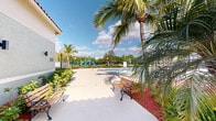 Located at 20950 NW 14th Pl in Miami Gardens, Florida, this single-level community amenity space offers a well-appointed outdoor environment designed for relaxation and recreation. At its centerpiece is a generously sized resort-style swimming pool with a smooth, light-colored deck that provides ample room for lounging, complemented by a separate spa for additional leisure. Tall, swaying palm trees and vibrant tropical plantings with red mulch landscaping beds frame the area, creating a lush South Florida atmosphere throughout. A shaded pergola structure near the pool offers a comfortable retreat from the sun, while classic wooden park benches are thoughtfully placed along the walkways for additional seating. A colorful children's playground with slides is situated at the far end of the grounds, making the space family-friendly. The main building features a distinctive Mediterranean-inspired architectural style, with a red barrel-tile roof and a soft sage-green exterior. Manicured shrubs and ornamental plants line the building's façade, enhancing its curb appeal. A gated entry provides security and privacy for the community. The entire outdoor space benefits from abundant natural sunlight and open sky views, embodying the vibrant and welcoming character of Miami Gardens living.