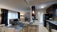 This single-floor 1-bedroom, 1-bathroom residence features an open-concept layout that seamlessly connects the kitchen and living areas, creating a cohesive and functional living environment. The kitchen is a standout feature, showcasing a granite-topped breakfast bar with seating for two, dark espresso cabinetry, and stainless steel appliances. A stylish pendant light hangs above the bar, adding a warm, focused glow that complements the overall aesthetic. The kitchen flows directly into the living area, which is brightened by a large window dressed with vertical blinds and dark curtain panels, allowing for flexible light control. The living space offers ample room for comfortable seating arrangements and is defined by neutral wall tones that complement the warm flooring underfoot. Architectural beam detailing on the ceiling adds subtle visual interest overhead. A hallway visible from the main living space leads to additional areas of the unit, maintaining a logical and accessible floor plan. The overall ambiance balances contemporary style with everyday practicality, highlighted by modern decor accents, cohesive color tones, and a layout designed to maximize the efficient use of space throughout.