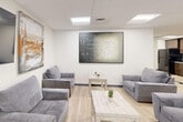Located at 2712 Santa Maria Way in Santa Maria, California, this single-floor space offers a well-organized and efficiently designed layout across three distinct areas. The interior features light wood-style flooring throughout, white walls, and a drop tile ceiling with recessed panel lighting that creates a bright, evenly lit environment.

The kitchen is a standout feature, outfitted with rich dark brown cabinetry that extends from floor to ceiling, providing ample storage. Black appliances — including a full-size refrigerator, electric range with oven, built-in microwave, and a range hood — are neatly integrated into the cabinetry. White countertops provide a clean contrast against the dark wood tones, and a sink is conveniently positioned at the end of the counter run.

The lounge areas are designed for comfort and social interaction. One area features a gray sofa paired with a wall-mounted flat-screen television and a white coffee table, accented by a framed abstract artwork. A second lounge space is more expansive, accommodating multiple gray sofas and armchairs arranged around light-toned coffee tables, with large abstract canvas paintings adorning the walls. The overall ambiance is calm and welcoming, with a cohesive neutral color palette that gives the space a clean, professional, yet comfortable feel.