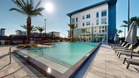 Located at 7000 Kiran Patel Dr in Winter Garden, Florida, this outdoor amenity space presents a resort-inspired environment designed for relaxation and social gathering. The centerpiece is a generously sized swimming pool with crystal-clear water, framed by elegant large-format stone tile decking that reflects the Florida sunshine throughout the day. Towering palm trees are strategically placed throughout the courtyard, lending a tropical ambiance that complements the warm Central Florida climate.

The surrounding multi-story building features a striking contemporary facade of white vertical paneling and expansive floor-to-ceiling glass, allowing interior spaces to connect visually with the outdoor amenity area. Along the poolside, lounge chairs offer ample space for sunbathing, while raised deck areas equipped with pergola structures provide shaded seating with stylish slatted overhead panels. These elevated lounges feature comfortable sectional seating, creating intimate gathering spots with landscaped planters and colorful flowering shrubs adding natural vibrancy.

An outdoor dining terrace with table-and-chair sets sits adjacent to the building's glass facade, ideal for al fresco meals. A dedicated grilling station completes the outdoor experience, making the space well-suited for social events. The overall design balances modern architecture with lush landscaping to create a cohesive, upscale outdoor living environment.