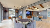Located at 1100 Warburton Rd in Summerville, SC, this two-story amenity clubhouse offers a generously proportioned layout spread across two floors, with the main level providing the majority of the functional space. The ground floor serves as the heart of the facility, anchored by an expansive, open-plan living area defined by rich wood beam accents, wide-plank light hardwood flooring, and floor-to-ceiling black-framed windows that flood the interior with natural light and provide views of the surrounding greenery and outdoor amenity areas. A statement fireplace wall with built-in shelving anchors one end of the main lounge, while comfortable seating arrangements invite gathering and relaxation. The dining room is a refined space, featuring sage-green accent walls, a dramatic sculptural chandelier, and a large communal table—ideal for private events or resident gatherings. A combined dining and living area, accessible through striking oak-framed glass sliding doors, opens to an intimate round-table setting bathed in natural light. A dedicated game room on the main level accommodates a billiards table and card table seating, bordered by expansive windows overlooking the courtyard. The upper floor houses additional lounge and hallway areas, a private office with a built-in desk and curated coastal artwork, and staircase connections to the main level. Throughout, the design blends contemporary and transitional aesthetics with warm, welcoming finishes.
