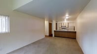 This single-floor space showcases an open-concept layout that seamlessly connects the living area to the kitchen, creating a cohesive and functional floor plan. The living area is finished with neutral-toned wall-to-wall carpeting, offering a comfortable and understated foundation for furnishings. Smooth, light-colored walls are complemented by a textured ceiling, and flush-mount ceiling fixtures provide consistent overhead lighting throughout the space.

The kitchen is positioned along one side of the open layout and features a raised peninsula with wood-toned cabinetry along its base, serving as a natural divider between the living and cooking areas. Upper cabinetry with lighter finishes lines the kitchen walls, providing ample storage. The kitchen includes a range with overhead ventilation, a refrigerator, and a sink, offering essential appliances for everyday use. A hallway visible from the main living space leads toward additional areas of the unit, suggesting a practical and well-organized floor plan. Overall, the space presents a clean, functional design with a warm, neutral palette throughout.
