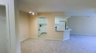 This single-floor space features an open-concept layout that seamlessly connects the living area to the kitchen. The living area is finished with wall-to-wall neutral carpet, contributing to a clean and understated aesthetic. Warm beige walls run throughout, creating a cohesive and inviting atmosphere. Track lighting is installed on the ceiling of the main living space, providing adjustable, functional illumination.

The kitchen is accessible via an open archway and is also visible through a pass-through breakfast bar, which features a curved white countertop edge — ideal for casual dining or food preparation. The kitchen is equipped with white cabinetry, a white electric range, a full-size refrigerator, and ample counter space. The cabinetry extends to the upper walls, maximizing storage. The flooring in the kitchen transitions from the carpeted living area to a hard surface, maintaining a practical distinction between spaces.

Overall, the layout is efficient and functional, with a bright, neutral palette that lends itself to easy personalization. The open sightlines between the kitchen and living area enhance the sense of spaciousness throughout the unit.