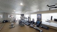 This fitness center offers a dedicated space for health and wellness, thoughtfully outfitted with a diverse selection of exercise equipment. The room features multiple treadmills, an elliptical machine, a stationary recumbent bike, and a multi-station cable weight machine, providing options for both cardio and strength training workouts. A wall-mounted flat-screen television is positioned for easy viewing during exercise sessions. The space is finished with a textured ceiling and is equipped with a ceiling fan, ensuring comfortable air circulation throughout. Large windows along the walls allow natural light to fill the room, creating a bright and energizing atmosphere. A full-length mirror on one wall enhances the sense of openness while allowing users to monitor their form. Motivational fitness artwork adorns the walls, adding character to the environment. The flooring is designed to withstand high-impact activity, making it practical for daily use. Recessed lighting supplements the natural light, ensuring the space remains well-lit at all hours. Overall, this fitness center is a functional and inviting facility suited for a wide range of fitness routines and goals.