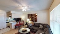 This single-floor space offers a well-appointed and comfortable layout designed for everyday living. The living area serves as the centerpiece of the home, featuring a large, plush sectional sofa arranged around a glass-topped coffee table, creating an inviting atmosphere for relaxing or entertaining. A decorative multi-panel wall art piece with an ornate scrollwork design in rich brown and gold tones adds a bold visual focal point to the room. A ceiling fan with an integrated light fixture is centrally mounted on the textured popcorn ceiling, providing both airflow and ambient lighting. Adjacent to the living area is a dedicated workspace or dining nook, outfitted with a desk or counter surface, a flat-screen monitor, and accented by red upholstered chairs, adding a pop of color to the neutral palette. Light-toned flooring runs throughout, complementing the soft wall colors and contributing to a bright, open feel. A table lamp on a side table adds warm, layered lighting to the space. The overall ambiance is warm and functional, balancing comfort with practical living amenities in a compact yet efficiently arranged floor plan.