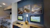 This single-floor space features a well-appointed resident services area designed for convenience and modern functionality. The focal point is a built-in workstation finished in a warm stone-toned countertop, housing multiple computer terminals that provide residents direct access to MyCamden.com for online rent payment and account management tasks.

The design aesthetic is contemporary and polished. A bold teal-blue accent wall serves as a striking backdrop, complementing the neutral tones of the surrounding walls and cabinetry. Three framed abstract art pieces with dynamic brushstroke patterns in gold, white, and dark tones are mounted prominently on the accent wall, adding visual interest and a curated, upscale feel to the space.

Overhead, sleek pendant lights hang at varying heights, providing focused task lighting over the workstation while contributing to the space's modern character. A decorative glass vase in amber tones is placed between the monitors, adding a refined finishing touch. The overall ambiance is clean, functional, and thoughtfully designed, reflecting a professional environment intended to serve residents efficiently.