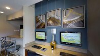 This single-floor space features a well-appointed resident services area designed for convenience and modern functionality. The focal point is a built-in workstation finished in a warm stone-toned countertop, housing multiple computer terminals that provide residents direct access to MyCamden.com for online rent payment and account management tasks.

The design aesthetic is contemporary and polished. A bold teal-blue accent wall serves as a striking backdrop, complementing the neutral tones of the surrounding walls and cabinetry. Three framed abstract art pieces with dynamic brushstroke patterns in gold, white, and dark tones are mounted prominently on the accent wall, adding visual interest and a curated, upscale feel to the space.

Overhead, sleek pendant lights hang at varying heights, providing focused task lighting over the workstation while contributing to the space's modern character. A decorative glass vase in amber tones is placed between the monitors, adding a refined finishing touch. The overall ambiance is clean, functional, and thoughtfully designed, reflecting a professional environment intended to serve residents efficiently.