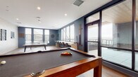 This single-floor amenity space is thoughtfully designed as an entertainment and social hub, combining a vibrant game room with comfortable lounge seating and direct access to an outdoor terrace. The layout centers around a full-size pool table with a rich wood frame and charcoal felt surface, complemented by a ping pong table that makes the most of the open floor plan. Warm hardwood-style flooring runs throughout, anchoring the space with a welcoming, contemporary feel.

One wall features a striking chevron-patterned tile accent, against which a large flat-screen TV is mounted alongside wall-mounted games including Connect Four and a chess board, creating an engaging recreational corner. A compact wet bar area adds convenience, showcasing sleek natural wood cabinetry with matte black hardware, a white countertop, and an artistic cloud-themed mural backdrop.

Large floor-to-ceiling windows bathe the room in natural light and frame expansive city skyline views, while a counter-height window bar lined with modern upholstered barstools offers an ideal perch to take in the scenery. Deep blue accent walls provide a bold contrast to the lighter tiled wall, giving the space a dynamic, modern character. Glass doors open directly onto an outdoor terrace, seamlessly extending the entertainment area and enhancing the elevated, airy ambiance throughout.