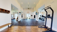 This expansive fitness center is thoughtfully designed to support a comprehensive workout experience across multiple dedicated training zones. Guests are welcomed through glass double doors into a bright entry area finished with warm hardwood-style flooring, a branded logo mat, built-in wall shelving, and seating benches — establishing a polished, community-focused first impression.

The main workout floor transitions to durable dark rubber tile flooring, purpose-built for high-intensity activity. Soaring ceilings with crown molding and recessed lighting lend an open, airy feel throughout, while multiple ceiling fans maintain comfortable airflow. Large windows and sliding glass doors line the perimeter, flooding the space with natural light and offering views of the surrounding landscape.

The strength training area is outfitted with a power rack, adjustable benches, a full dumbbell rack, kettlebells, medicine balls, and weight plates. A dedicated functional training zone includes a heavy punching bag, TRX-style suspension trainers, and plyo boxes. The cardio section features machines including a stair climber and rowing equipment. Wall-mounted flat-screen TVs are positioned throughout for entertainment during workouts.

Bold motivational graphics — including phrases like "Hustle & Muscle," "Harder. Better. Faster. Stronger." and "If it doesn't challenge you, it won't change you" — energize the space and reinforce its dynamic fitness-forward identity.