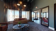 This single-floor community clubhouse is defined by its striking contemporary design and thoughtfully curated interiors. The welcoming lobby immediately impresses with soaring ceilings, dramatic floor-to-ceiling textured wall art in rich earth tones, and an abundance of natural light streaming through large picture windows that frame outdoor views. Large-format dark tile flooring anchors the entryway, while plush area rugs and a comfortable neutral sofa with accent pillows create an inviting seating arrangement beneath pendant lighting. The open lounge and recreation area transitions seamlessly from the lobby, showcasing a spacious layout with a billiard table as a central focal point, vibrant statement pendant lights in gold and amber tones, and bold geometric accent walls in yellow and charcoal. Built-in banquette seating with teal upholstery and an abstract art panel offer cozy nooks for casual gatherings, complemented by mixed flooring of tile and patterned carpet. Glass-partitioned office or meeting spaces with black steel-framed doors provide privacy without sacrificing visual openness. A resident mailbox area is conveniently integrated into the layout. Throughout, the design balances bold color accents with neutral backdrops, creating a modern, energetic, and welcoming communal environment.