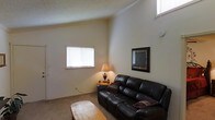 This single-story space features a living area defined by its notably high vaulted ceilings, which lend an open, airy quality to the room. The walls are finished in a soft, neutral cream tone that complements the textured ceiling, contributing to a warm and understated aesthetic throughout.

The living area is furnished with a dark leather reclining sofa accented by decorative pillows, paired with a rustic wooden coffee table that adds a natural, earthy element to the space. A framed wall mirror with a dark border serves as a focal point on one wall, enhancing the sense of depth in the room. A classic table lamp on a small side table provides warm ambient lighting, supplementing the natural light that enters through a window fitted with horizontal blinds.

The flooring is carpeted in a neutral tone, adding to the overall comfort of the space. A second, higher window near the ceiling allows additional natural light to filter in, further emphasizing the height of the vaulted ceiling. Overall, the living area conveys a cozy, relaxed atmosphere well-suited for everyday living.