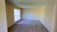 This single-story space features a generously sized main living area finished with wall-to-wall carpeting in a neutral gray-beige tone, offering a clean and versatile foundation for a variety of furniture arrangements. The walls are painted in a soft, warm off-white/light yellow hue that contributes to an open and airy atmosphere throughout the room. A large window fitted with vertical blinds allows ample natural light to filter into the space, brightening the interior while offering privacy control. The textured ceiling adds a subtle architectural detail consistent with the overall practical and functional design of the space. White baseboards provide a clean contrast against the carpeted floors and painted walls. The room's layout is straightforward and open, lending itself well to customization. Overall, the space presents a simple, move-in-ready environment with a calm, neutral aesthetic suited to a wide range of uses.