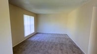 This single-story space features a generously sized main living area finished with wall-to-wall carpeting in a neutral gray-beige tone, offering a clean and versatile foundation for a variety of furniture arrangements. The walls are painted in a soft, warm off-white/light yellow hue that contributes to an open and airy atmosphere throughout the room. A large window fitted with vertical blinds allows ample natural light to filter into the space, brightening the interior while offering privacy control. The textured ceiling adds a subtle architectural detail consistent with the overall practical and functional design of the space. White baseboards provide a clean contrast against the carpeted floors and painted walls. The room's layout is straightforward and open, lending itself well to customization. Overall, the space presents a simple, move-in-ready environment with a calm, neutral aesthetic suited to a wide range of uses.