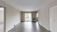 This single-floor residence offers a clean, functional layout spread across several well-defined living spaces. The main living area is bright and open, featuring smooth gray laminate flooring, neutral-toned walls, and generous natural light streaming in through a large sliding glass door that looks out onto a tree-lined outdoor setting. A wall-mounted air conditioning unit and baseboard heating are present, providing year-round climate control. An adjacent area, also finished with laminate flooring, offers additional living or dining space and features built-in louvered closet doors, adding practical storage. The kitchen is efficiently designed with dark cabinetry, a gas range, and stainless steel appliances including a refrigerator, set against dark hardwood-style flooring that adds a contemporary contrast. The bedrooms are carpeted for comfort and offer ample closet storage, with one room featuring a full wall of louvered bi-fold closet doors and a window overlooking the surrounding greenery. The bathroom is finished in clean white tile, complete with a full soaking tub and shower combination, built-in wall niches, and a frosted window for natural light and privacy. Overall, the space presents a tidy, neutral palette throughout.