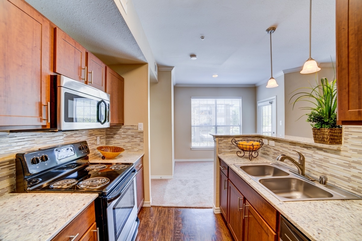 Cocina con gabinetes de castaño, encimeras de granito moteado, protector contra salpicaduras de azulejos, electrodomésticos de acero inoxidable y pisos de superficie dura. - eaves North Dallas