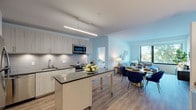 This single-floor residence showcases a thoughtfully designed open-concept layout that seamlessly connects the kitchen, dining, and living areas. The kitchen is well-appointed with light wood cabinetry extending to the ceiling, stainless steel appliances including a refrigerator, electric range, and microwave, along with a subway tile backsplash and dark quartz countertops. A central island provides additional workspace and casual dining space. Light wood-style flooring flows throughout the main living areas, enhancing the bright, airy atmosphere.

The living room benefits from a large picture window framing outdoor views of surrounding greenery, filling the space with natural light. The open dining area connects naturally to both the kitchen and living room, making the layout ideal for entertaining.

The primary bedroom features its own en-suite bathroom with a wide vanity, dark countertops, ample cabinetry, and direct access to a walk-in closet. A second bedroom offers comfortable proportions with wood-style flooring. A secondary bathroom includes a full tub-shower combination with clean white tile surround. Throughout the residence, the neutral color palette and modern finishes create a cohesive, contemporary ambiance.