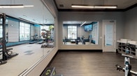 Located at 5500 Home Valley Dr in Raleigh, NC, this well-appointed fitness center offers a comprehensive workout environment designed for both functionality and aesthetic appeal. The space is spread across a single floor and is divided into distinct zones that cater to a variety of exercise needs.

The main workout area features a bold, contemporary design highlighted by teal accent walls, neutral tones, and a striking checkered tile floor. Overhead, linear pendant lighting and a ceiling fan ensure the space is well-lit and comfortable. Large windows with horizontal blinds line the walls, allowing ample natural light to fill the room while providing views of the surrounding exterior.

Cardio equipment, including treadmills and elliptical machines, is thoughtfully arranged in a dedicated section near the windows, offering natural light during workouts. The strength training zone houses a power rack, adjustable bench, dumbbells, kettlebells, medicine balls, and a multi-functional cable machine, providing a thorough free-weight and resistance training experience.

A separate studio room, enclosed with floor-to-ceiling glass walls and a large pass-through window, opens visually to the main floor. This room features hardwood-style flooring, wall-mounted TVs, and stationary bikes, making it ideal for cycling classes or focused training sessions. Wall-mounted TVs throughout the facility enhance the overall workout experience.