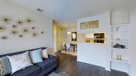 This single-floor residence offers a cohesive, open-concept design that seamlessly connects the main living areas. The living room provides a welcoming atmosphere, anchored by rich, dark hardwood flooring that extends throughout the space. Decorative wall accents, including a starburst wall piece, add a modern, curated touch to the neutral-toned walls accented by a textured ceiling overhead.

The living area flows naturally into the dining space, which is defined by a stylish pendant light suspended above a round dining table with contemporary chairs, set atop a plush area rug. This intimate dining nook creates a warm, functional area for everyday meals or gatherings.

The kitchen is positioned adjacent to the dining area and features a pass-through bar-height counter that maintains the open feel of the floor plan. Light cabinetry lines the upper shelving, complemented by black appliances including a microwave and range. A bold, gold decorative wall panel adds an elegant focal point above the kitchen area.

A hallway visible from the main living space leads to additional rooms, offering clear separation between the shared living areas and private spaces. Throughout the residence, the combination of warm wood tones, neutral walls, and thoughtfully chosen decor creates a balanced, modern ambiance.