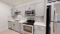 This single-floor space showcases a thoughtfully designed, modern interior with a seamless open-concept layout connecting the kitchen and living areas. Light hardwood flooring flows consistently throughout, lending warmth and continuity to the space.

The kitchen is a standout feature, equipped with a full suite of stainless steel appliances — including a refrigerator, range, microwave, and dishwasher — complemented by sleek gray shaker-style cabinetry and crisp white quartz countertops. A peninsula with an undermount sink serves as both a functional workspace and a natural divider between the kitchen and living area. Striking black pendant lights hang above the peninsula, adding a modern, design-forward touch. A built-in wine rack provides an additional stylish storage accent.

The living area is bright and open, benefiting from multiple windows fitted with horizontal blinds that allow natural light to filter in. A ceiling fan overhead ensures year-round comfort. An exterior door provides direct access to the outdoors.

The bedroom is generously proportioned, with light-filled windows, a ceiling fan, and the same light hardwood flooring that carries throughout the unit.

The bathroom features a white quartz vanity with gray cabinetry, a large framed mirror, and a soaking tub with tiled surround, completing this clean and contemporary residence.