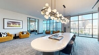 Located in the heart of Houston at 808 Crawford St, this elegantly designed amenity space occupies a single floor and offers an impressive blend of modern aesthetics and functional versatility. The open layout encompasses a combined dining and living area alongside a dedicated private dining room, all unified by a cohesive contemporary design language.

Floor-to-ceiling black-framed glass walls and windows are a defining architectural feature throughout, bathing the interior in abundant natural light while framing sweeping views of the Houston downtown skyline and an adjoining landscaped terrace. The main gathering area centers around a striking oval white table suspended beneath a dramatic sculptural chandelier of brass and globe pendants, complemented by plush upholstered seating. Bold amber lounge chairs positioned near the expansive windows create an inviting informal seating nook with unobstructed city views.

The private dining room is distinguished by a dark wood conference-style table, warm pendant lighting, and bold contemporary artwork, with direct access to the outdoor terrace through tall glass doors. A refined media wall clad in vertical wood slat paneling houses built-in dark cabinetry and a mounted flat-screen display, adding warmth and sophistication. Textured blue-grey carpeting and curated art pieces complete the polished, upscale ambiance throughout the space.
