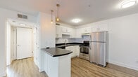 This thoughtfully designed 1-bedroom, 1-bathroom unit offers a functional and modern layout across a single floor. Light wood-look flooring runs consistently throughout, lending warmth and visual continuity to every room.

The kitchen is a standout feature, outfitted with crisp white shaker cabinetry, dark countertops, and a full suite of stainless steel appliances including a refrigerator, electric range, and oven. A peninsula with a curved dark countertop provides additional workspace and casual seating, while two Edison-style pendant lights add a stylish, contemporary touch overhead.

The living room is bright and open, featuring a ceiling fan with light fixture and glass double doors that invite natural light and provide direct access to an outdoor area. The neutral white walls create a clean, versatile backdrop throughout the space.

The bedroom is generously sized, featuring a window with natural views, a dedicated closet with wire shelving for practical storage, and direct access to the hallway leading to adjacent rooms.

The bathroom is finished in a clean, minimal style with wood-look flooring, a white vanity with drawer storage, a full soaking tub and shower combination with brushed nickel fixtures, and a wall-mounted sconce for focused lighting. The overall ambiance of the unit is fresh, modern, and move-in ready.