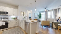 This well-designed single-floor residence offers a bright, open-concept layout that seamlessly connects the kitchen, dining, and living areas. The kitchen is a standout feature, equipped with white shaker-style cabinetry, granite countertops, stainless steel appliances including a side-by-side refrigerator, range, and built-in microwave, and a central island with pendant lighting overhead and a stainless steel sink. Light wood-style flooring runs throughout the main living areas, enhancing the airy, contemporary feel. The living room is generously sized, filled with natural light from large windows with plantation shutters, and features a ceiling fan for added comfort. Multiple bedrooms are present throughout the residence, each carpeted and outfitted with ceiling fans and windows that bring in ample natural light. The primary bedroom includes access to a private en-suite bathroom featuring a walk-in shower with tile surround, a granite-topped vanity with white cabinetry, and wood-style flooring. A secondary bathroom mirrors the refined finishes with a granite vanity, tub-shower combination, and tile surround. The overall ambiance throughout is clean, modern, and cohesive, with a neutral color palette that creates a welcoming and functional living environment.